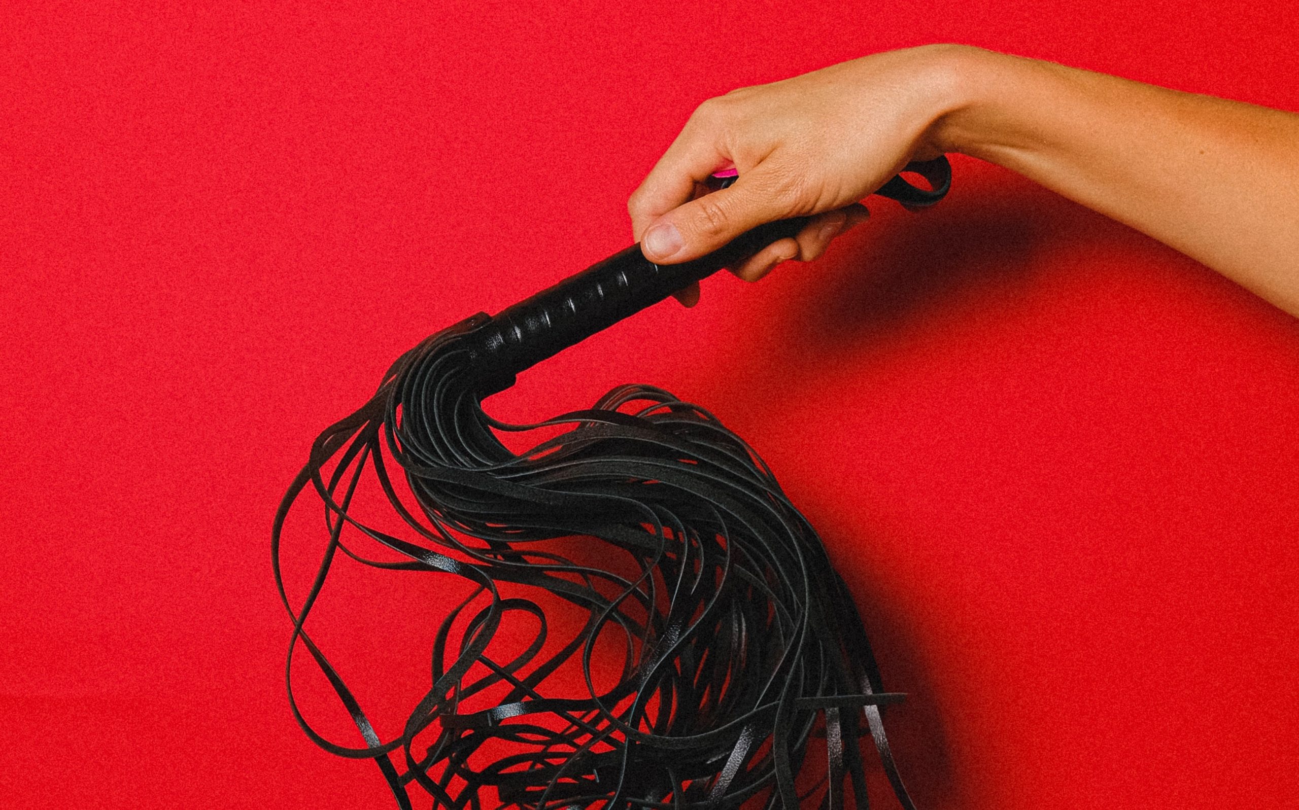 Imagem de uma mão segurando um chicote de couro preto com várias tiras sob um fundo vermelho. Ilustrando Fantasias e Fetiches